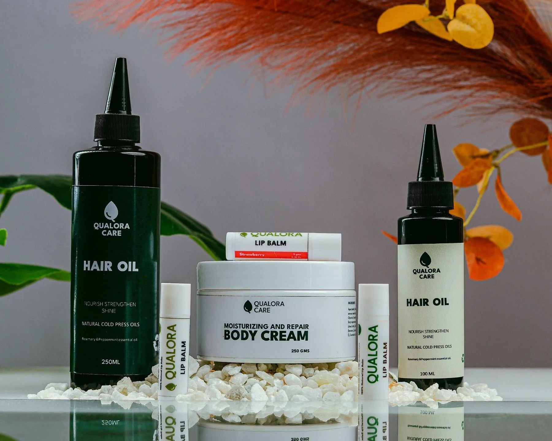 The Qualora Care collection: Hair Oil 250 ml and 100 ml, Body Cream and Lip Balms on white stones with pampas and tropical leaves